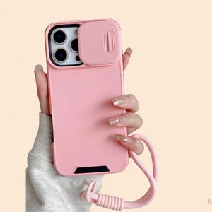 Puede incluir: Una funda de teléfono rosa con una cubierta para la lente de la cámara y un cordón rosa a juego. La funda tiene un acabado liso y un acento negro en la parte inferior. La funda está diseñada para proteger el teléfono.