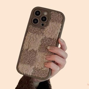 Könnte beinhalten: Eine braun-beige karierte Handyhülle mit flauschiger Textur. Die Hülle hat einen braunen Rand und einen Kameraausschnitt. Die Handyhülle wird in einer Hand gehalten.