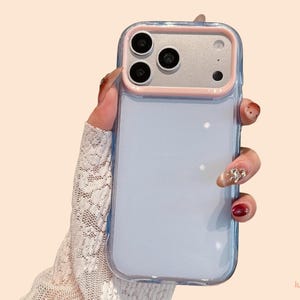 May include: A clear phone case with a light blue back and a pink camera border. The case has a transparent border and is designed to fit a smartphone. The phone case is held by a person.