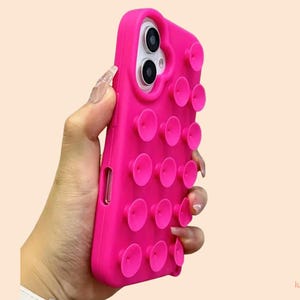Könnte beinhalten: Eine leuchtend pinke Handyhülle mit mehreren Saugnapf-ähnlichen Aufsätzen. Die Hülle ist für ein Smartphone konzipiert, mit Aussparungen für Kamera und Tasten. Die Hülle wird in einer Hand vor einem hellpfirsichfarbenen Hintergrund gehalten.
