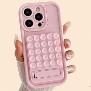 May include: A light pink phone case with a pop-it style design and a kickstand. The case has a smooth finish and is designed to fit a smartphone. The case has a camera cutout and a logo in the bottom right corner.