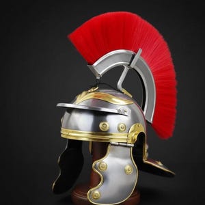 Może przedstawiać: Srebrny hełm rzymski ze złotymi akcentami i żywym czerwonym pióropuszem. Hełm posiada wizjer, osłony policzkowe i ozdobne nity. Ta historyczna replika jest prezentowana na drewnianym stojaku na ciemnym tle.