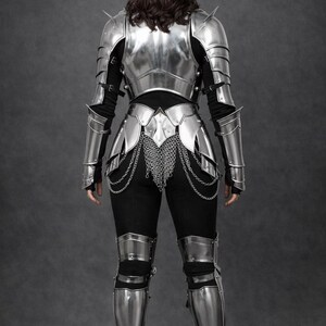 May include: A full suit of polished silver armor, including a breastplate, shoulder guards, arm and leg protection, and a chainmail skirt. The armor is worn over a black long-sleeved shirt and pants. The image is taken from the back.