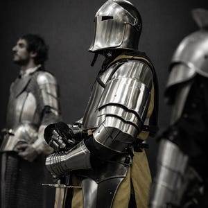 Puede incluir: Un caballero medieval con armadura de acero pulido, que incluye casco, peto y protectores de brazos. El caballero sostiene una espada, con una t&uacute;nica amarilla visible debajo de la armadura. La imagen es en escala de grises.