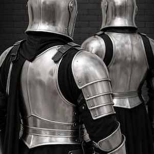 May include: Two sets of medieval armor, including helmets and chest plates, are displayed against a dark brick wall. The armor is made of shiny, silver-colored metal. The helmets have a pointed top.