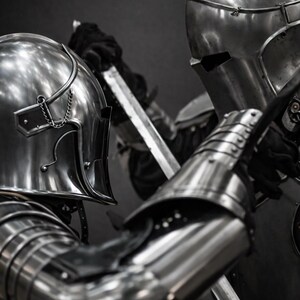 Puede incluir: Dos figuras con armadura de acero pulido, involucradas en una escena de combate medieval. Los cascos y la armadura articulada reflejan la luz, resaltando los detalles de la artesan&iacute;a. Las espadas est&aacute;n levantadas, sugiriendo un momento de conflicto.