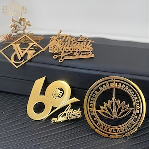 May include: Gold-colored metal pins and brooches with various designs. One pin features the text "The Sunset Box.confture" with scissors and a comb. Another displays the number "60" with the text "Años" and "2°AGYM Osormo".