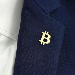 May include: A gold-colored Bitcoin symbol pin is affixed to a navy blue blazer. The pin is positioned on the lapel, near a white collared shirt. The Bitcoin symbol is a stylized letter "B" with vertical lines.