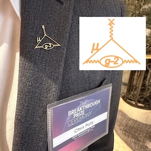 May include: A gold-colored pin featuring a diagram with the Greek letter mu, the text "g-2", and a wavy line design. The pin is attached to a dark gray blazer. A clear plastic holder with text is in the pocket.