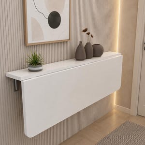 May include: A white wall-mounted folding table with a smooth rectangular surface. On top of the table sits a small green potted plant and three dark gray decorative vases of varying shapes. The table is attached to a vertical slatted wall panel.
