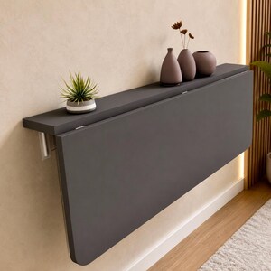 May include: A dark grey wall-mounted folding table with a smooth rectangular surface. On the top shelf sits a small potted succulent plant in a grey and white pot, alongside three smooth, rounded decorative vases in shades of brown and grey.