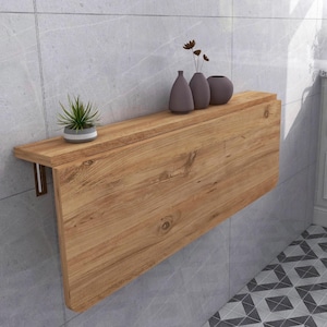 Könnte beinhalten: Ein an der Wand montierter, klappbarer Holztisch mit natürlicher Holzmaserung. Der Tisch wird von einer Metallhalterung getragen und hat eine kleine Topfpflanze und drei dekorative Vasen. Der Tisch ist etwa 76 cm lang.