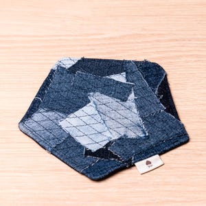 Könnte beinhalten: Ein Denim-Untersetzer mit Patchwork-Design. Der Untersetzer besteht aus verschiedenen Blautönen von Denim, die in einem geometrischen Muster zusammengenäht sind. Ein kleines Etikett mit einem Logo ist an der Seite angebracht. Der Untersetzer liegt auf einer hellen Holzoberfläche.