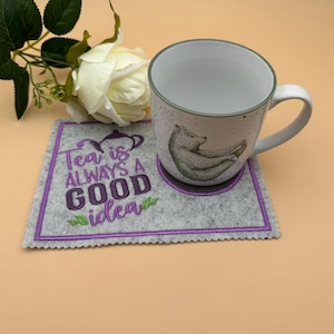 Könnte beinhalten: Eine weiße Keramiktasse mit einer Bärenillustration, die auf einem grauen Filzuntersetzer mit dem Text "Tea is ALWAYS A GOOD idea" in Lila steht. Eine weiße Rose und grüne Blätter befinden sich im Hintergrund.