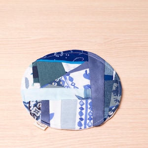 May include: A round, patchwork coaster featuring a variety of blue fabrics. The coaster is made from different shades of blue, including light blue, navy, and patterned fabrics. The coaster is a handmade item.