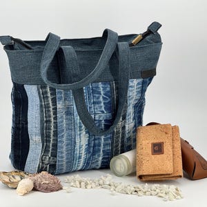 Könnte beinhalten: Eine Tragetasche aus vertikalen Streifen von blauem Jeansstoff in verschiedenen Schattierungen mit zwei passenden Henkeln und einem Reißverschluss. Daneben liegen eine beigefarbene Korkgeldbörse, ein braunes Brillenetui und eine weiße zylindrische Flasche.