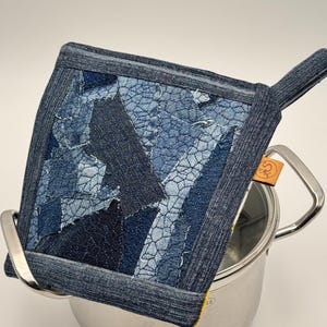Op de afbeelding: Een vierkante pannenlap met een patchworkontwerp van diverse blauwe spijkerstofresten met zichtbare stiksels, een donkere spijkerstof rand en een ophanglus. Een klein bruin leren label met een rond logo is aan de zijkant bevestigd.