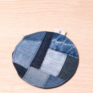 May include: A round coaster made from repurposed denim patches in various shades of blue. The coaster has a patchwork design and a small tag attached. A sustainable and stylish home decor item.