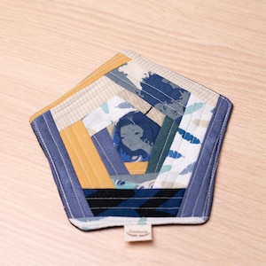 May include: A pentagon-shaped fabric coaster with a patchwork design. The coaster features a variety of fabrics in blue, yellow, and beige tones, with a central image of a woman's face. A small tag is attached.