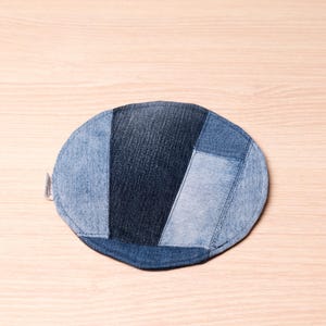 Może przedstawiać: Okrągły podkładka pod szklankę z denimu wykonana z przetworzonych dżinsów. Podkładka ma wzór patchworkowy z różnych odcieni niebieskiego denimu. Mała metka jest przymocowana z boku. Podkładka ma średnicę około 10 cm.