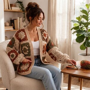 Könnte beinhalten: Eine langärmelige, vorne offene Strickjacke aus gehäkelten Granny Squares in den Farben Creme, Olivgrün und Rostorange. Das Kleidungsstück hat einen V-Ausschnitt, gerippte Kanten und zwei Vordertaschen und wird über einem weißen Tanktop getragen.
