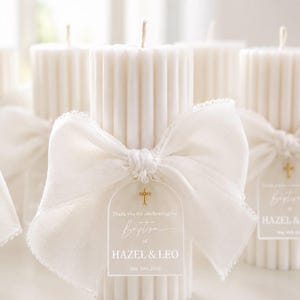 May include: A white fluted pillar candle adorned with a large white fabric bow and a translucent arched tag. The tag features a small gold cross icon and the printed text: Thank you for celebrating the Baptism of HAZEL & LEO May 30th, 2026.