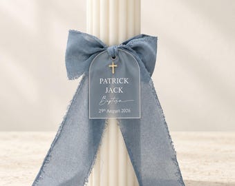 Puede incluir: Una vela de pilar blanca acanalada adornada con un lazo de cinta azul polvoriento deshilachado y una etiqueta acrílica semicircular. La etiqueta presenta un icono de cruz dorada y el texto impreso: PATRICK JACK Baptism 29th August 2026.