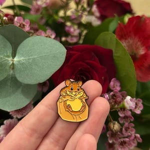 May include: A gold-colored enamel pin featuring a cartoon hamster holding a large, round object. The pin has a black outline and is set against a blurred background of red roses and other flowers.