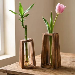 May include: Two wooden vases with glass tubes, each holding a plant. One vase contains a green bamboo stalk, and the other holds a pink tulip. The vases are on a wooden table near a window.
