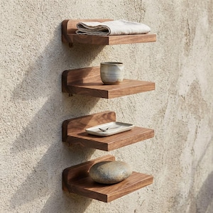 Op de afbeelding: Vier houten zwevende planken aan een getextureerde muur. Elke plank bevat een item: een gevouwen doek, een keramische beker, een klein bord met een lepel en een gladde steen. De planken zijn warmbruin.
