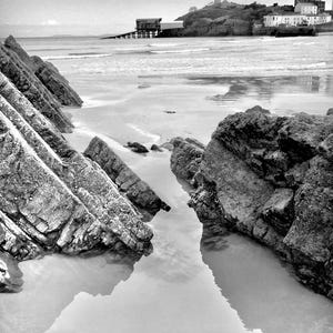 May include: Monochrome image of a coastal landscape. Rugged, layered rocks lead the eye towards a sandy beach and the sea. A pier and buildings are visible on a distant island, under a cloudy sky, with the water reflecting the sky.