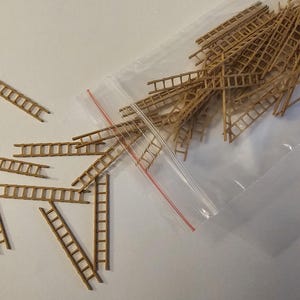 Può includere: Una collezione di piccole scale in legno color marrone chiaro, alcune sparse e altre all'interno di un sacchetto di plastica trasparente. Le scale sono in miniatura, con pioli e supporti laterali visibili, suggerendo un tema artigianale o di modellismo.