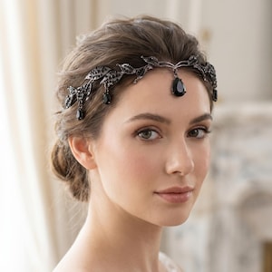 Op de afbeelding: Een donker metalen bruids hoofdband met zwarte druppelvormige accenten. De hoofdband heeft een delicaat, bladachtig ontwerp met bungelende zwarte edelstenen. De vrouw op de foto draagt de hoofdband.