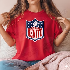 Può includere: T-shirt rosso con un design a scudo con un pallone da football e stelle. Il testo "TEAM BENITO" è visualizzato in un carattere in grassetto in stile collegiale. La maglietta è un classico girocollo.