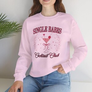 May include: Pale pink sweatshirt with burgundy text reading "SINGLE BABES Cocktail Club". The design includes a cocktail glass with a cherry, surrounded by leaves and hearts. The model is wearing blue jeans.