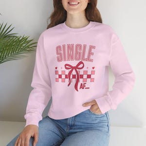 Peut inclure: Un sweat-shirt rose clair avec le texte "SINGLE AF..." dans un design rouge et rose. Le motif comprend un nœud et un motif à carreaux. Le sweat-shirt est à manches longues et en matière douce.