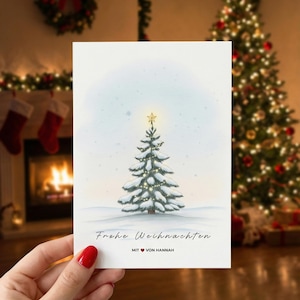 May include: A Christmas card featuring a watercolor illustration of a snow-covered Christmas tree with a star on top. The card has the text "Frohe Weihnachten" and "MIT VON HANNAH". The background shows a fireplace and Christmas decorations.