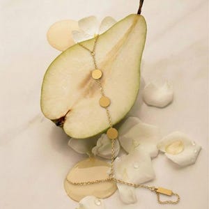 May include: A gold-colored hand chain with circular and rectangular charms is draped over a sliced pear. The pear is green with a brown stem, and the background features white flower petals and a light-colored surface.