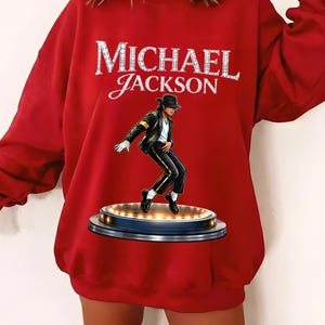 May include: A red sweatshirt featuring the name "MICHAEL JACKSON" in a sparkling, silver font. Below the text is an illustration of Michael Jackson in a signature pose, wearing a black suit and hat, standing on a stage.