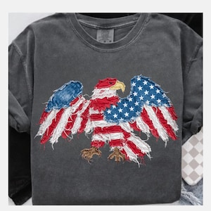 May include: Dark gray t-shirt featuring a distressed American flag eagle design. The eagle's wings are made of the US flag with red and white stripes and blue star fields. The eagle's beak is yellow and the claws are brown.