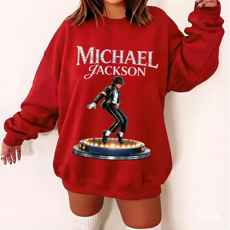 May include: A red sweatshirt featuring the name "MICHAEL JACKSON" in a sparkling, silver font. Below the text is an illustration of Michael Jackson in a signature pose, wearing a black suit and hat, standing on a stage.