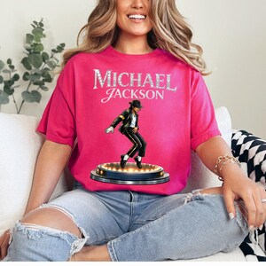 May include: A hot pink t-shirt featuring the name "MICHAEL JACKSON" in a sparkling, silver font. Below the text is an illustration of Michael Jackson in a black suit, white shirt, and black hat, performing on a stage with lights.