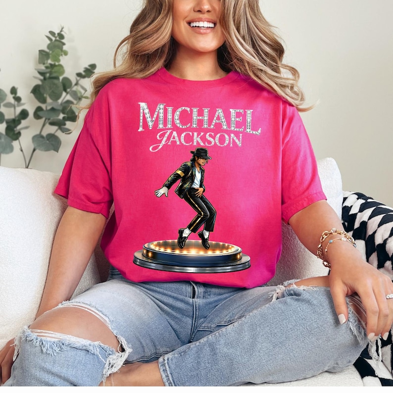 May include: A hot pink t-shirt featuring the name "MICHAEL JACKSON" in a sparkling, silver font. Below the text is an illustration of Michael Jackson in a black suit, white shirt, and black hat, performing on a stage with lights.