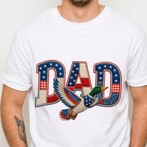 Könnte beinhalten: Weißes T-Shirt mit patriotischem Design. Das Wort "DAD" ist in Rot, Weiß und Blau mit Sternen und Streifen gehalten. Unter dem Wort ist eine fliegende Ente in ähnlichen patriotischen Farben gestickt.