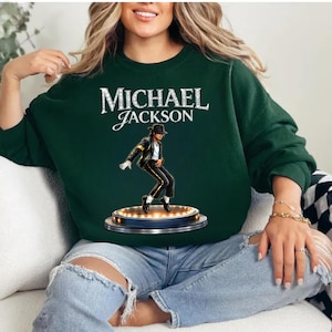 May include: Dark green sweatshirt featuring the text "MICHAEL JACKSON" in white script. Below the text is an illustration of Michael Jackson in a black suit and hat, performing on a stage with lights. The sweatshirt is a solid color.