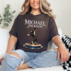 May include: A brown t-shirt featuring the name "MICHAEL JACKSON" in a sparkling font. Below the text is an image of Michael Jackson in a black outfit, striking a dance pose on a stage with lights. The shirt is a casual wear item.
