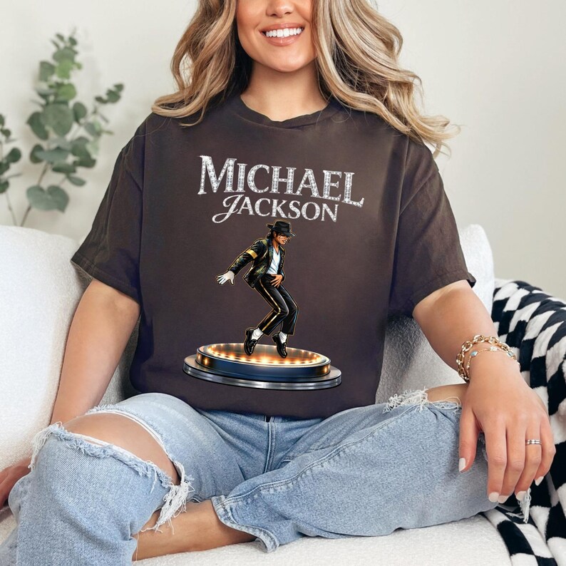 May include: A brown t-shirt featuring the name "MICHAEL JACKSON" in a sparkling font. Below the text is an image of Michael Jackson in a black outfit, striking a dance pose on a stage with lights. The shirt is a casual wear item.