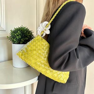 May include: A woven, lime green handbag with a braided strap and a white flower accent. The bag has a rectangular shape and a textured surface. The strap is a lighter shade of green.