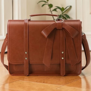 May include: A brown leather satchel with a large bow detail. The bag has a top handle and a shoulder strap. The front flap is secured with two straps and metal hardware. The bag is approximately 12 inches wide and 10 inches tall.