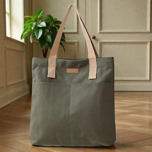Può includere: Una borsa tote in tela verde con due tasche frontali e manici beige. La borsa ha una piccola toppa rettangolare beige. La borsa tote è appoggiata su un pavimento in legno.